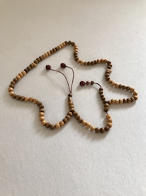 A neatly arranged string of wooden prayer beads lies on a light-colored fabric surface. The beads vary slightly in shade, creating an organic pattern, with a tassel attached to the end. The overall layout forms a looped, abstract shape.