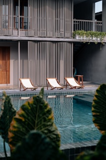 Three lounge chairs with wooden frames and white cushions sit by a swimming pool, in front of a modern building with vertical slatted design elements. Lush green plants border the foreground, while the pool reflects the surrounding architecture.