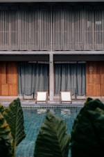 Two white lounge chairs are placed beside a tranquil swimming pool, with large leafy plants in the foreground. The background features a modern building with vertical wooden slats covering the upper floor and large wooden doors on the lower level.