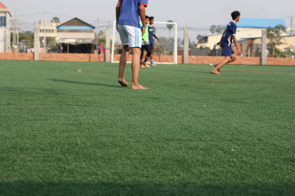 A group of young people are playing soccer on a green artificial turf field. The focus is on a few players in sportswear, with some of them appearing to be barefoot. A goalpost is visible in the background, along with a surrounding wire fence. Buildings and trees can be seen in the distance beyond the field.