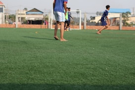 A group of young people are playing soccer on a green artificial turf field. The focus is on a few players in sportswear, with some of them appearing to be barefoot. A goalpost is visible in the background, along with a surrounding wire fence. Buildings and trees can be seen in the distance beyond the field.