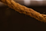 A close-up view of a thick, twisted brown rope set against a dark background. The rope's fibrous texture is clearly visible, highlighting its coarse and rugged appearance.