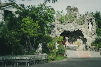 A peaceful outdoor religious grotto is surrounded by lush green trees and vegetation. At the center, there is a statue of a religious figure inside a rocky cave, with another statue on the right side of the cave entrance. Colorful flowers are placed at the base of the statue, adding vibrant touches to the scene. Benches are arranged in front of the grotto for visitors or worshippers to sit. The atmosphere is serene and contemplative.