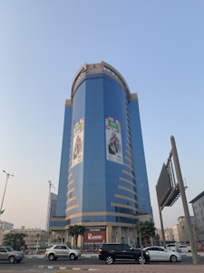 A tall, modern skyscraper dominates the scene with a blue and gold facade. Large promotional banners are displayed on the building's exterior. Several cars are seen driving and parked along the street in front of it. The sky is clear with a slight gradient of blue, and palm trees line the sidewalk.