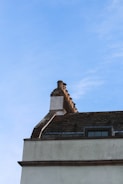 A rooftop with a row of decorative brick chimney pots set against a clear blue sky. The roof is slanted and covered in wooden or slate shingles. Part of a window is also visible beneath the roofline.
