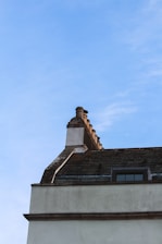 A rooftop with a row of decorative brick chimney pots set against a clear blue sky. The roof is slanted and covered in wooden or slate shingles. Part of a window is also visible beneath the roofline.