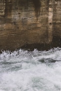 A textured, weathered concrete wall is partially submerged in churning, frothy water. The water appears to be rapidly moving and splashes against the wall, creating a dynamic contrast between the man-made surface and the natural element.