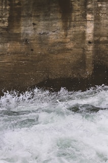A textured, weathered concrete wall is partially submerged in churning, frothy water. The water appears to be rapidly moving and splashes against the wall, creating a dynamic contrast between the man-made surface and the natural element.