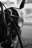 Close-up view of a bicycle, focusing on the handlebars, brake cables, and headlamp. The image is in black and white, giving it a classic, timeless feel. The background is blurred, highlighting the foreground details like the shiny metal parts and the texture of the cables.