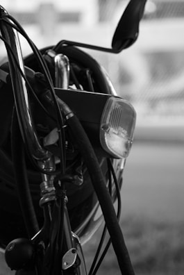 Close-up view of a bicycle, focusing on the handlebars, brake cables, and headlamp. The image is in black and white, giving it a classic, timeless feel. The background is blurred, highlighting the foreground details like the shiny metal parts and the texture of the cables.
