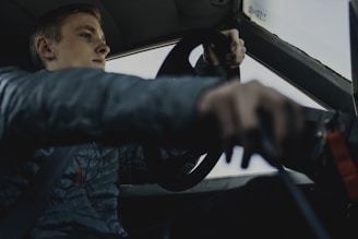 A person is seated in a car, gripping the steering wheel tightly while looking out the windshield with focus. The individual is wearing a dark jacket and appears to be driving.