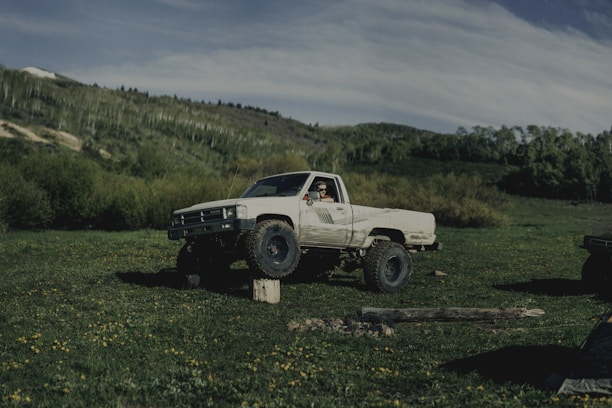 A large white pickup truck is parked off-road in a lush green field surrounded by dense trees and hills. The truck has oversized tires and is positioned with one wheel elevated on a small tree stump. A person is visible in the driver's seat, wearing sunglasses and looking outwards. The sky is partly cloudy, creating a serene and adventurous atmosphere.