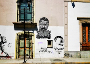 A city street scene features a building facade with artistic graffiti. The artwork includes a detailed monochrome portrait of a man with a stern expression. Below the portrait are the words 'ASARO acosadores.' To the left, there is an image of a person walking and an anarchist symbol. A streetlamp stands in front of the building, and the windows have ornate grilles.