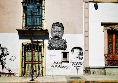 A city street scene features a building facade with artistic graffiti. The artwork includes a detailed monochrome portrait of a man with a stern expression. Below the portrait are the words 'ASARO acosadores.' To the left, there is an image of a person walking and an anarchist symbol. A streetlamp stands in front of the building, and the windows have ornate grilles.