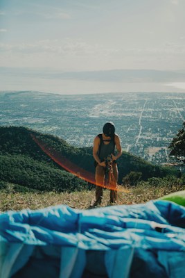 A person is preparing for paragliding on a grassy hill overlooking a cityscape and distant body of water. The person is wearing a hat and handling the lines connected to a blue and green parachute laid out on the ground.