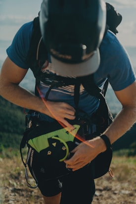 A person wearing a helmet and a blue shirt is seen adjusting a strap on a backpack. The scene is outdoors, possibly in a mountainous or hilly area, with a blurred background of natural landscape.