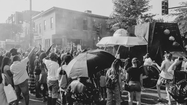 A large crowd of people gathers on a city street, engaging with a parade or public event. Several individuals hold umbrellas, while others raise their hands in the air, indicating enthusiasm or celebration. A float shaped like a spacecraft or UFO is part of the procession, adorned with planetary models, and a person is standing on it. The scene is lively, with a mix of adults and children, framed by buildings and trees in the background.