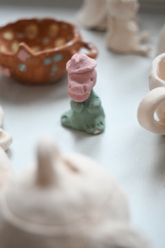 A small, artistic clay figure painted in pastel colors, primarily pink and mint. It is surrounded by other blurred ceramic objects, including a patterned, orange pottery dish with blue and yellow spots.