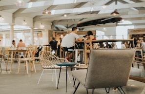 A bright and spacious cafe or food court environment with people sitting and enjoying their meals. The setting features various types of seating, including wicker chairs and cushioned armchairs. The ceiling is high with visible beams, and there are several hanging lights and open spaces. Several tables are filled with patrons engaged in conversation.