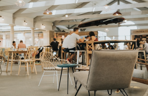 A bright and spacious cafe or food court environment with people sitting and enjoying their meals. The setting features various types of seating, including wicker chairs and cushioned armchairs. The ceiling is high with visible beams, and there are several hanging lights and open spaces. Several tables are filled with patrons engaged in conversation.