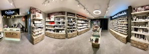 A well-organized chocolate shop interior with multiple shelves displaying various chocolate products. The shop is brightly lit, with modern fixtures and a clean design. There is a counter on the left with a display and a sign reading 'Hotel Chocolat'. The shelves are stocked with neatly arranged boxes, jars, and bottles of chocolates and related products. The layout is spacious, allowing for easy browsing.