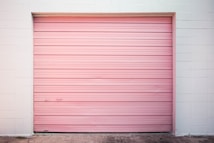 A closed pink rolling garage door set within a white brick wall. The surface is smooth with horizontal slats and minimal detailing. The ground is concrete.