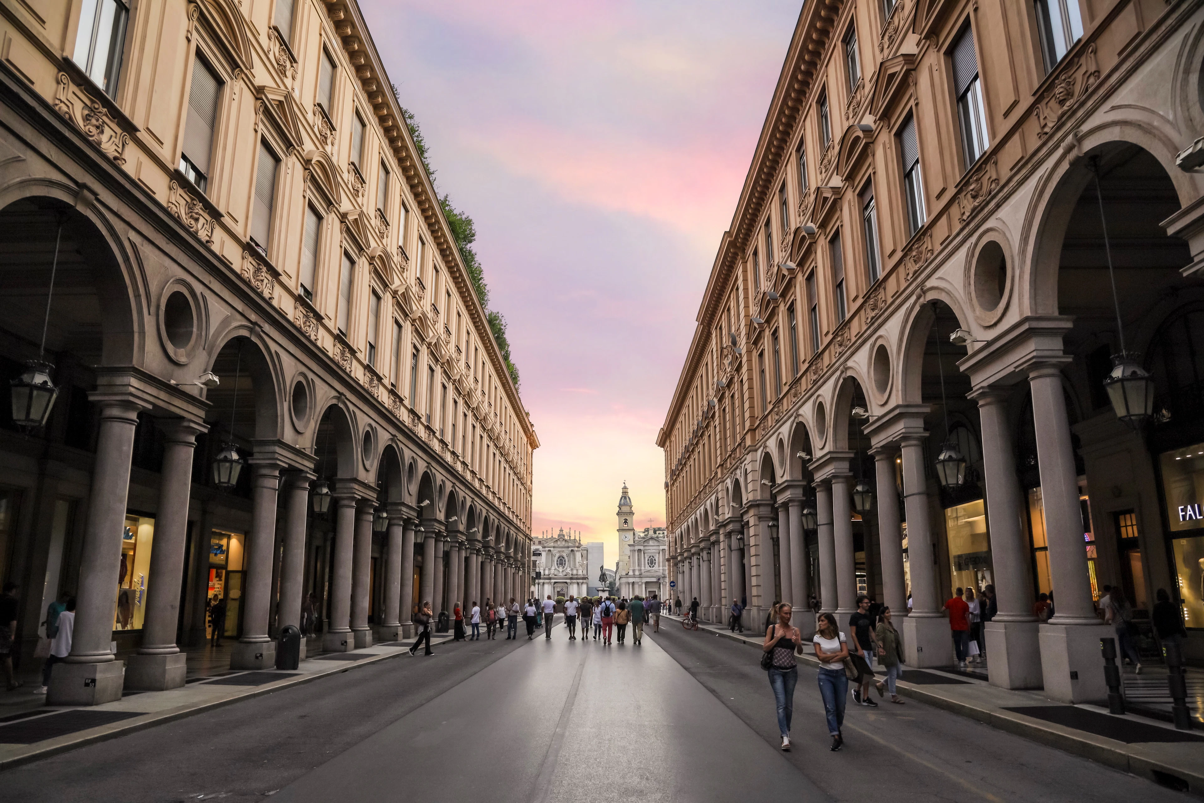 Via Roma in Turin at dusk, its long colonnaded arcades receding on both sides of the empty street under a pink and lilac sky, with a tower lit at the far end