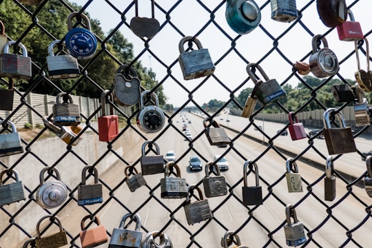 A chain-link fence is covered with various padlocks, including combination locks and solid metal locks, hanging in front of a highway with several cars driving by. The background includes trees and a concrete barrier.