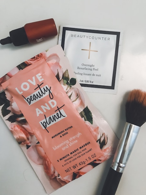 A collection of skincare and beauty products laid out on a surface. The items include a sachet labeled 'Love Beauty and Planet' featuring a floral design with pink flowers, a small dropper bottle with a dark cap, a white packet labeled 'Beautycounter Overnight Resurfacing Peel,' and a makeup brush with dark bristles and a metallic handle.