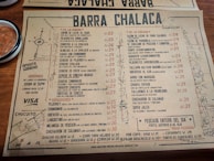 A rustic paper menu from a restaurant called Barra Chalaca, featuring a variety of dishes with prices. The menu is divided into sections like 'De La Barra', 'De La Cocina', and 'Sánguches', each with a list of items. Side notes on the left indicate opening hours and location. The background includes a simple map sketch, and a bowl with red sauce is visible at the top left corner.