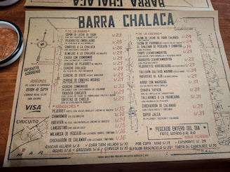 A rustic paper menu from a restaurant called Barra Chalaca, featuring a variety of dishes with prices. The menu is divided into sections like 'De La Barra', 'De La Cocina', and 'Sánguches', each with a list of items. Side notes on the left indicate opening hours and location. The background includes a simple map sketch, and a bowl with red sauce is visible at the top left corner.