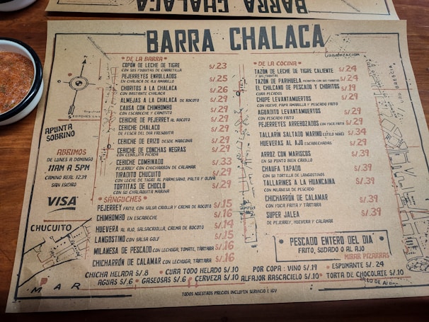 A rustic paper menu from a restaurant called Barra Chalaca, featuring a variety of dishes with prices. The menu is divided into sections like 'De La Barra', 'De La Cocina', and 'Sánguches', each with a list of items. Side notes on the left indicate opening hours and location. The background includes a simple map sketch, and a bowl with red sauce is visible at the top left corner.