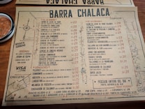 A rustic paper menu from a restaurant called Barra Chalaca, featuring a variety of dishes with prices. The menu is divided into sections like 'De La Barra', 'De La Cocina', and 'Sánguches', each with a list of items. Side notes on the left indicate opening hours and location. The background includes a simple map sketch, and a bowl with red sauce is visible at the top left corner.