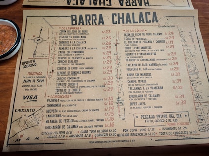 A rustic paper menu from a restaurant called Barra Chalaca, featuring a variety of dishes with prices. The menu is divided into sections like 'De La Barra', 'De La Cocina', and 'Sánguches', each with a list of items. Side notes on the left indicate opening hours and location. The background includes a simple map sketch, and a bowl with red sauce is visible at the top left corner.