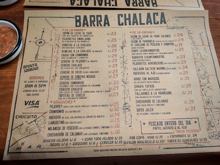 A rustic paper menu from a restaurant called Barra Chalaca, featuring a variety of dishes with prices. The menu is divided into sections like 'De La Barra', 'De La Cocina', and 'Sánguches', each with a list of items. Side notes on the left indicate opening hours and location. The background includes a simple map sketch, and a bowl with red sauce is visible at the top left corner.