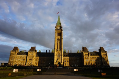 A large, historic government building with Gothic architecture stands prominently under a partly cloudy sky. The building features a tall central clock tower with a flag on top, surrounded by symmetrical wings. The façade is illuminated by warm, golden sunlight, highlighting the intricate stone detailing and gothic spires.