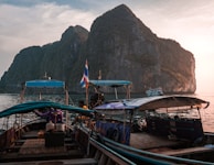 Several traditional long-tail boats are docked on a tranquil body of water, with towering, rocky cliffs in the background. The sky is partly cloudy and the sun appears to be setting, casting a warm glow on the scene. A Thai flag is visible on one of the boats, and there are some people aboard, seemingly preparing or resting.