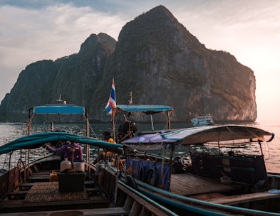 Several traditional long-tail boats are docked on a tranquil body of water, with towering, rocky cliffs in the background. The sky is partly cloudy and the sun appears to be setting, casting a warm glow on the scene. A Thai flag is visible on one of the boats, and there are some people aboard, seemingly preparing or resting.