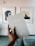 A hand is holding a journal titled 'My Life Journal' against the backdrop of a stylish interior. The journal cover is a muted grey with a minimalistic design. Two framed abstract artworks are hung on a pale wall. The hand has neatly manicured nails, and the room exudes a sense of calm and modern aesthetics.