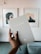 A hand is holding a journal titled 'My Life Journal' against the backdrop of a stylish interior. The journal cover is a muted grey with a minimalistic design. Two framed abstract artworks are hung on a pale wall. The hand has neatly manicured nails, and the room exudes a sense of calm and modern aesthetics.