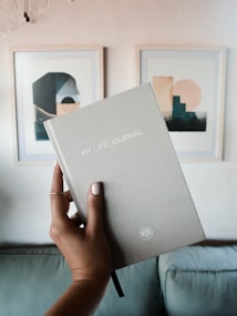 A hand is holding a journal titled 'My Life Journal' against the backdrop of a stylish interior. The journal cover is a muted grey with a minimalistic design. Two framed abstract artworks are hung on a pale wall. The hand has neatly manicured nails, and the room exudes a sense of calm and modern aesthetics.