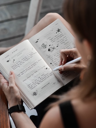 A person is writing in a journal or planner, holding a pen in their right hand. The pages are decorated with small illustrations of fish and starfish, along with handwritten notes. The notebook appears open and rests on the person's lap, with their left hand supporting it. The background shows a wooden floor and part of a wooden table.