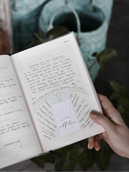 A hand holding a notebook or journal open to a page with handwritten notes and a motivational insert that says 'RADIATE self-love'. The page includes words and phrases related to self-love and personal growth. A blue and green woven bag is visible in the background.