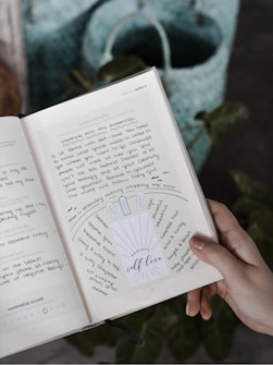 A hand holding a notebook or journal open to a page with handwritten notes and a motivational insert that says 'RADIATE self-love'. The page includes words and phrases related to self-love and personal growth. A blue and green woven bag is visible in the background.