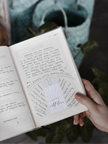 A hand holding a notebook or journal open to a page with handwritten notes and a motivational insert that says 'RADIATE self-love'. The page includes words and phrases related to self-love and personal growth. A blue and green woven bag is visible in the background.