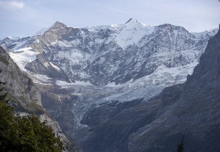 A stunning mountain landscape featuring snow-capped peaks and a rugged, rocky terrain. The scene is accentuated by patches of ice and snow that cascade down the slopes, contrasting with the stark, dark rock faces. Sparse vegetation can be seen at the lower elevations, bringing a touch of green to the otherwise monochromatic scene.