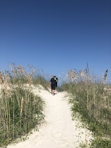 A mentor and a young man walking together along a sandy path lined with soft purple wildflowers.