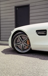 A close-up of a white car showcasing its front wheel and part of the side panel. The wheel has a detailed, modern design, and the panel displays the 'V8 BITURBO' badge. The background includes a gray building wall with a black door numbered '2'.