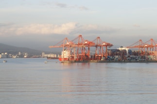 A port area with large red cranes positioned along the shoreline, used for loading and unloading containers from ships at a dock. Several stacked shipping containers are visible, and smaller boats are in the water nearby. The landscape in the background includes distant city buildings and hills under a partly cloudy sky.