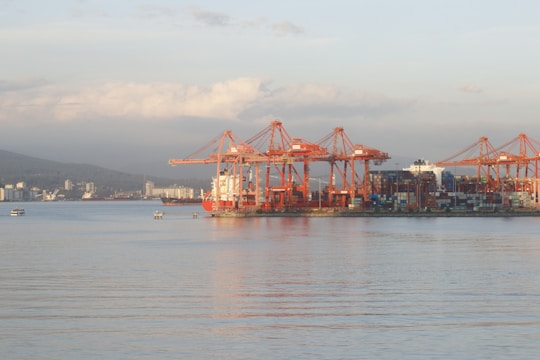 A port area with large red cranes positioned along the shoreline, used for loading and unloading containers from ships at a dock. Several stacked shipping containers are visible, and smaller boats are in the water nearby. The landscape in the background includes distant city buildings and hills under a partly cloudy sky.